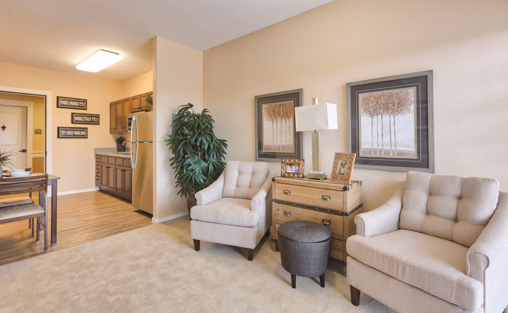 A cozy senior living area featuring two beige armchairs with tufted backs, a wooden chest used as a side table with a lamp and framed photos on top, and two framed tree paintings on the wall. To the left, there is a dining table with chairs and a kitchen area with wooden cabinets and a stainless steel refrigerator. The room has beige walls and carpeted flooring with wood flooring in the kitchen area.