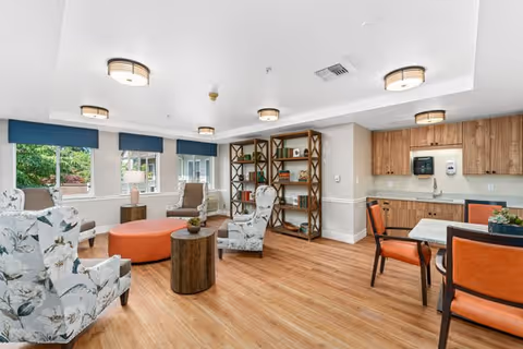 A bright and spacious common area in a senior living facility featuring floral upholstered armchairs, an orange round ottoman, wooden side tables, a bookshelf with decorative items, a small kitchenette with wooden cabinets, and a dining table with orange cushioned chairs. Large windows with blue valances allow natural light to fill the room.