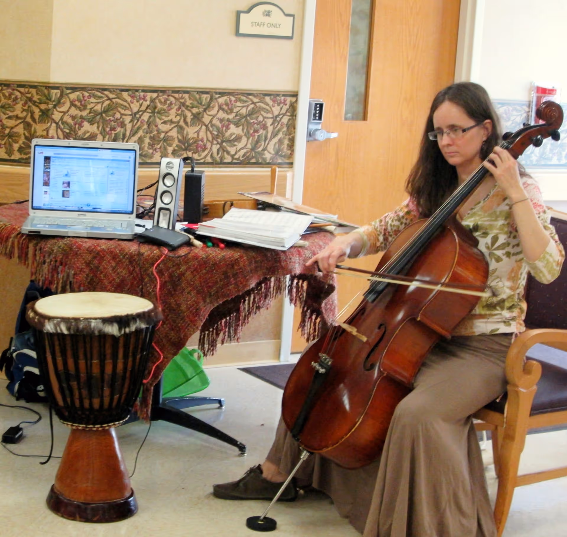 A woman seated on a wooden chair playing a cello in a room with a table covered by a red patterned cloth. On the table, there is a laptop, speakers, and some papers. A drum is placed on the floor next to the table. A door with a 'Staff Only' sign is visible in the background.