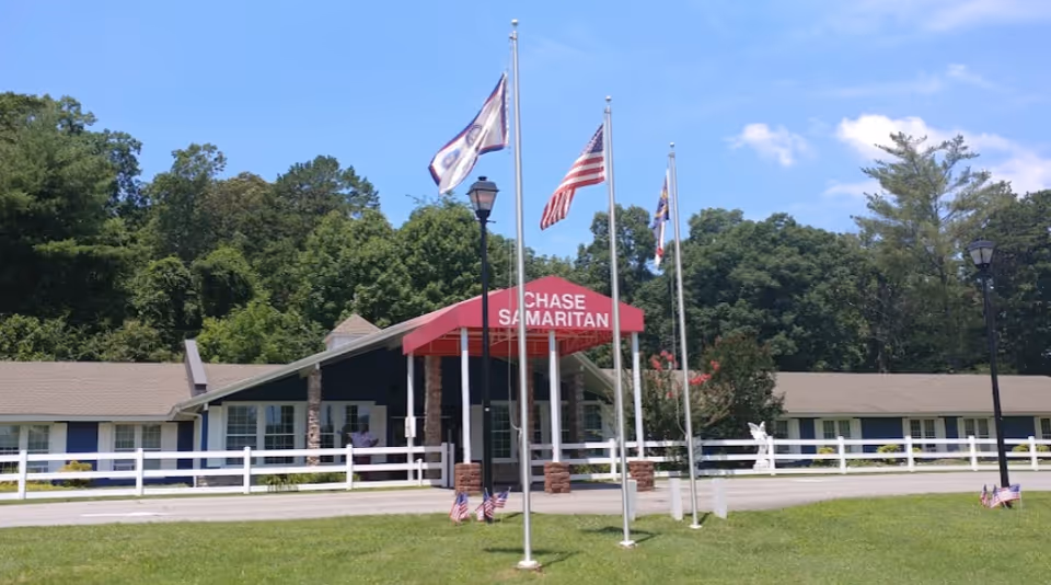 Exterior view of a single-story building with a red awning that reads 'CHASE SAMARITAN'. The building is surrounded by a white fence and green lawn with several flagpoles displaying flags, including the American flag. Trees and a clear blue sky are visible in the background.