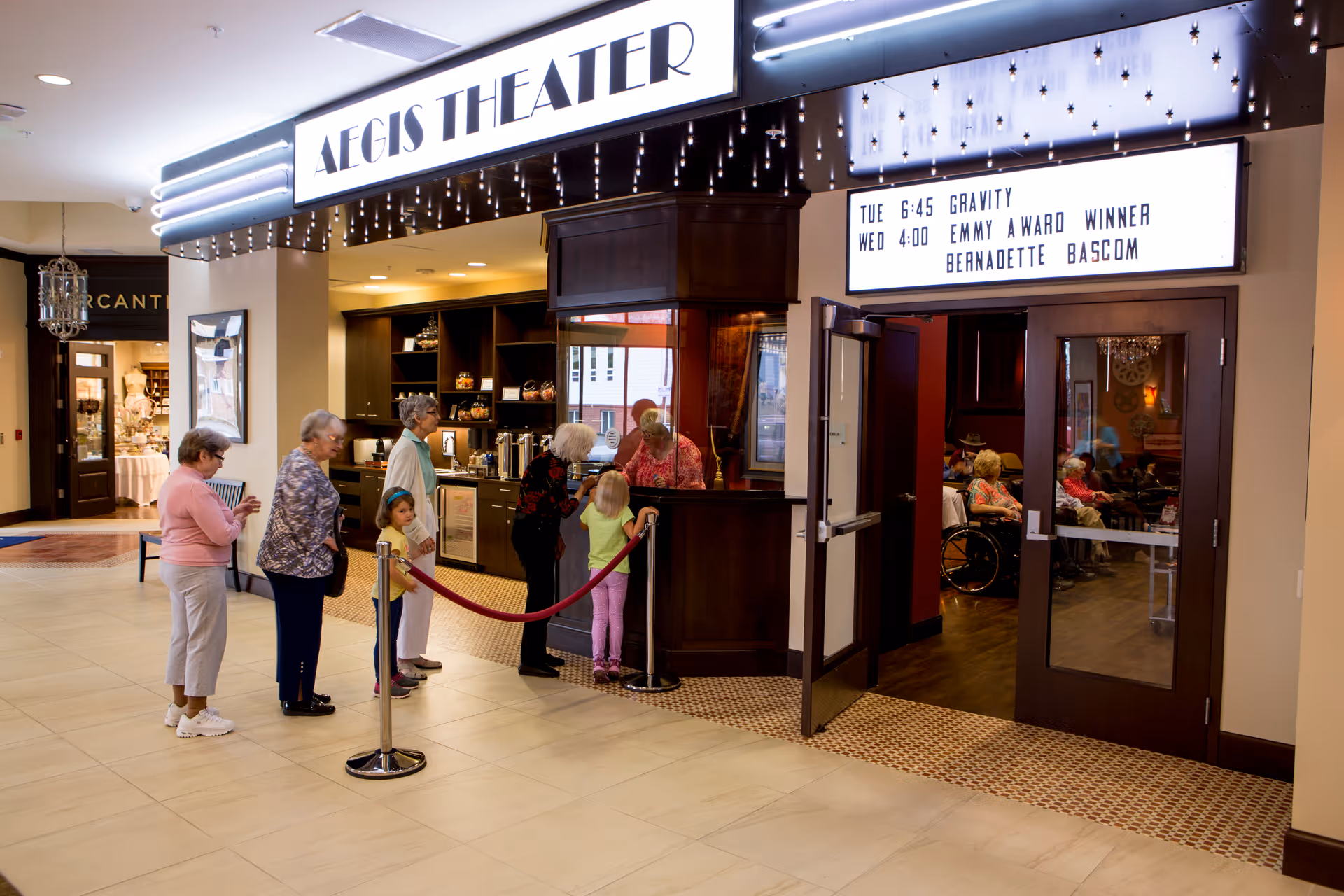 A group of elderly people and two children standing in line at the ticket counter of Aegis Theater inside a senior living facility. The theater entrance has a marquee displaying movie times and events. Inside the theater, several elderly individuals are seated, some in wheelchairs.