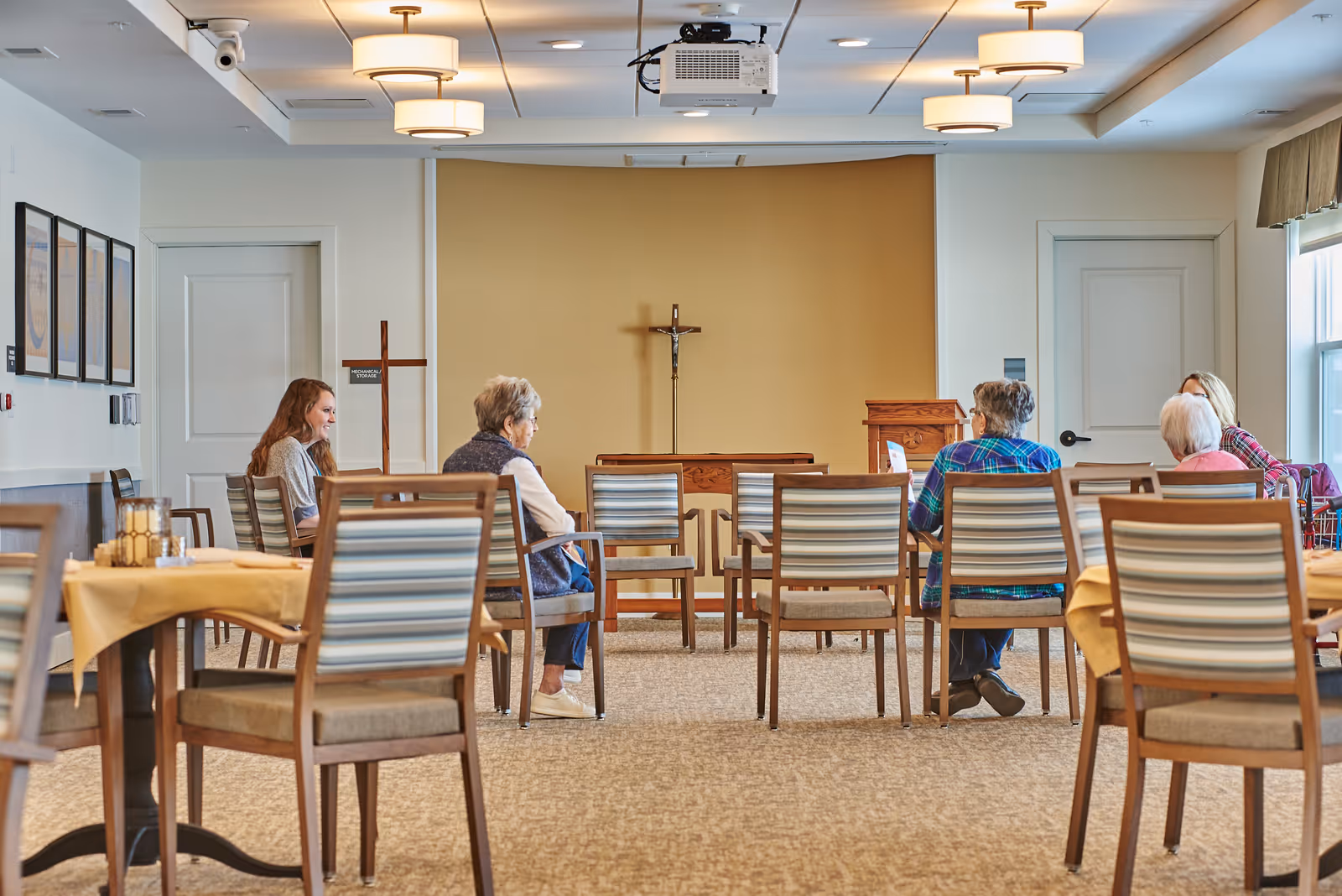 A small group of elderly people seated on chairs in a room with a cross and a podium at the front, suggesting a chapel or meeting space within a senior living community. The room has beige walls, carpeted floor, and ceiling lights, with tables covered in yellow tablecloths on the sides.