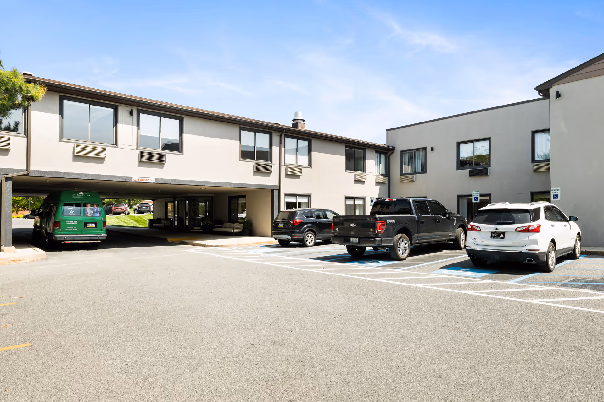 Exterior view of Willow Brook Rehabilitation and Healthcare Center showing a two-story building with several windows, a covered driveway area, and a parking lot with multiple vehicles including a green van and several cars parked in designated spaces.