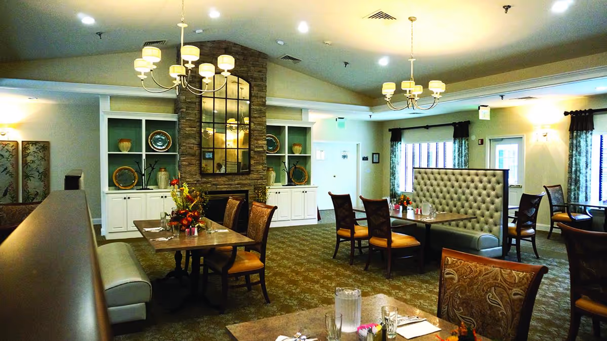 A spacious dining room with multiple tables and chairs arranged neatly. The room features a stone fireplace with a large mirror above it, flanked by built-in white cabinets displaying decorative plates and vases. There are chandeliers hanging from the ceiling, and windows with curtains allowing natural light to enter. The carpeted floor and comfortable seating create a warm and inviting atmosphere.