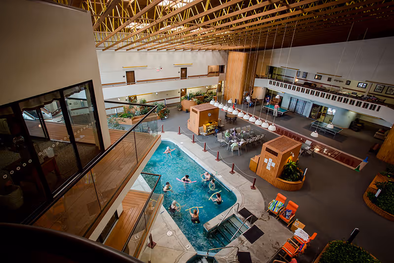 Indoor recreational area in a senior living facility featuring a small swimming pool with several people exercising in the water. Surrounding the pool are lounge chairs with towels, tables and chairs for seating, and a ping pong table. The area has a high ceiling with wooden beams and a second-floor balcony overlooking the space.