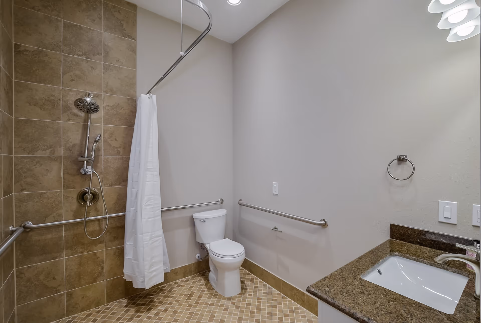 A clean and accessible bathroom featuring a tiled walk-in shower with a white shower curtain, a white toilet with grab bars on both sides, and a sink with a granite countertop and a mirror above it.