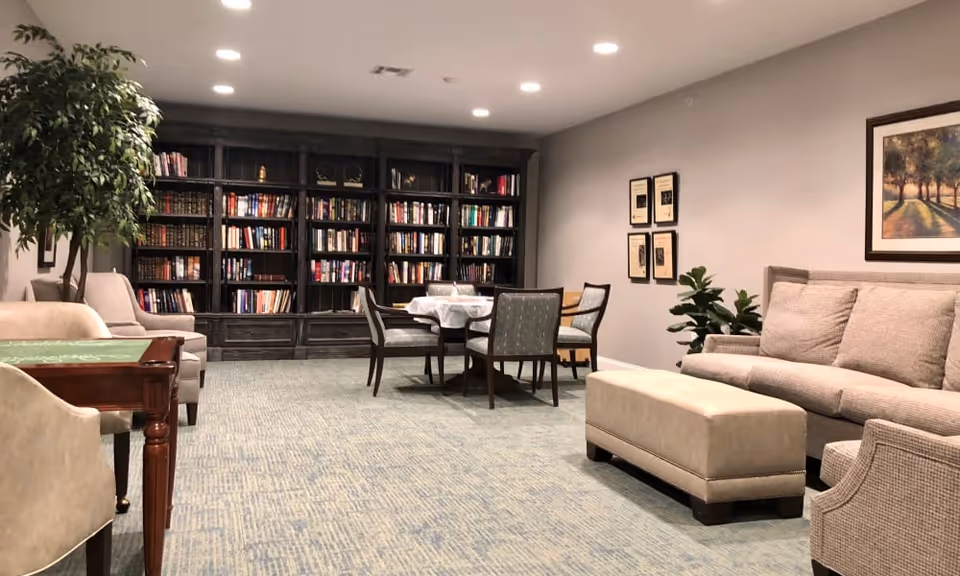 Cozy communal lounge with large bookshelves, a table and chairs, sofa and ottoman, and potted plants.