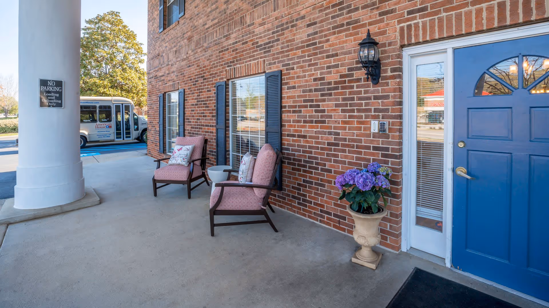 Covered entrance area of a brick building with two cushioned chairs and a small table between them, a potted plant with purple flowers, a blue door with a window, and a white column with a no parking sign. A shuttle bus is visible in the background.