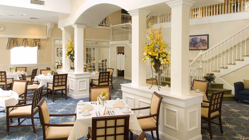 A bright and elegant dining room with multiple tables covered in white tablecloths, each set with napkins, cups, and silverware. The room features beige walls, blue patterned carpet, and decorative columns with large floral arrangements. A staircase with white railings is visible in the background along with a seating area.