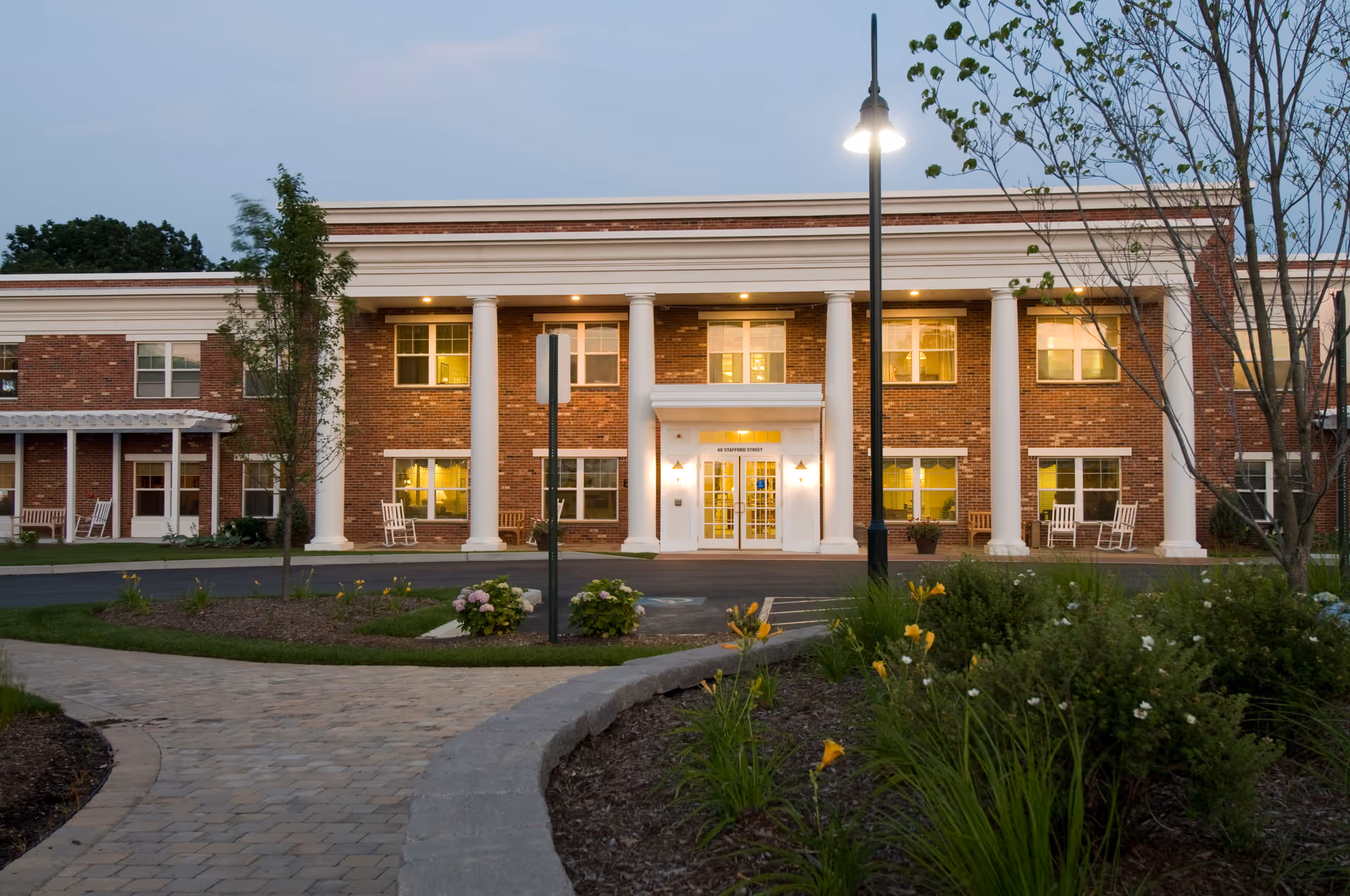 Front exterior of a two-story brick senior living building with white columns, an illuminated entrance, rocking chairs on the porch, a paved walkway and landscaped grounds at dusk.