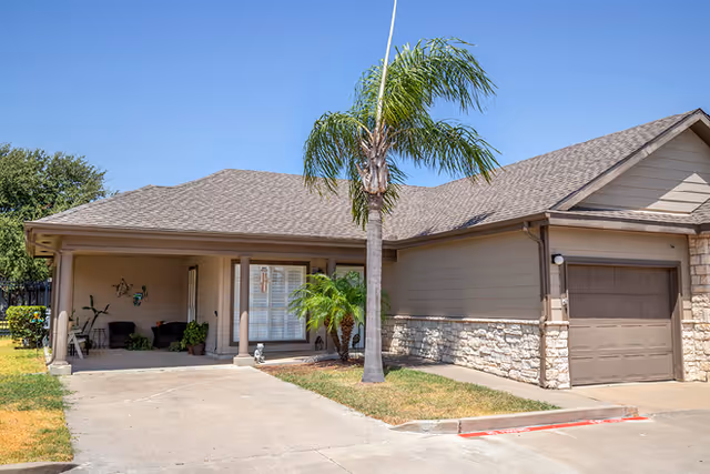 Single-story residential-style building front with a covered entrance, attached garage, palm tree, and driveway.