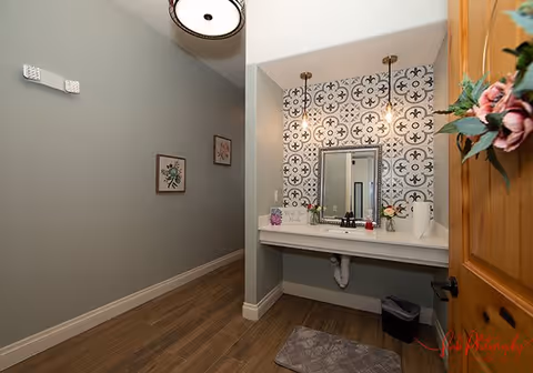 Vanity sink area with patterned tile backsplash, mirror, pendant lights and a hallway with wood-look flooring.