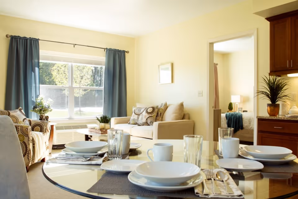 A bright living room with a large window covered by blue curtains, a beige sofa with patterned cushions, and a glass dining table set with white plates, bowls, cups, and glasses. A doorway leads to a bedroom with a bed, lamp, and small plant visible. Wooden kitchen cabinets and a potted plant are on the right side.