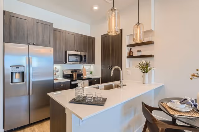 Modern kitchen with dark wood cabinets, stainless steel refrigerator, microwave, and stove. A white countertop island with a sink and two pendant lights above it. A tray with glassware is on the island, and a small dining table with place settings is visible on the right.