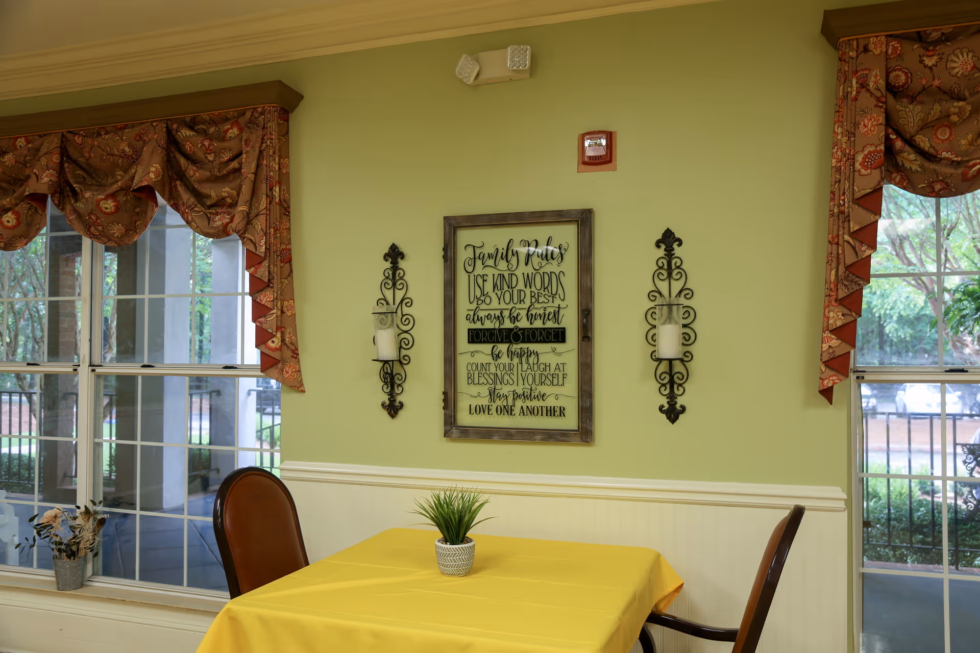 A small dining area with a table covered in a yellow tablecloth and two chairs. On the table is a small potted plant. The wall behind the table is light green with white wainscoting and features a framed sign with family rules and two decorative candle holders. Two windows with patterned valances show an outdoor view with greenery.