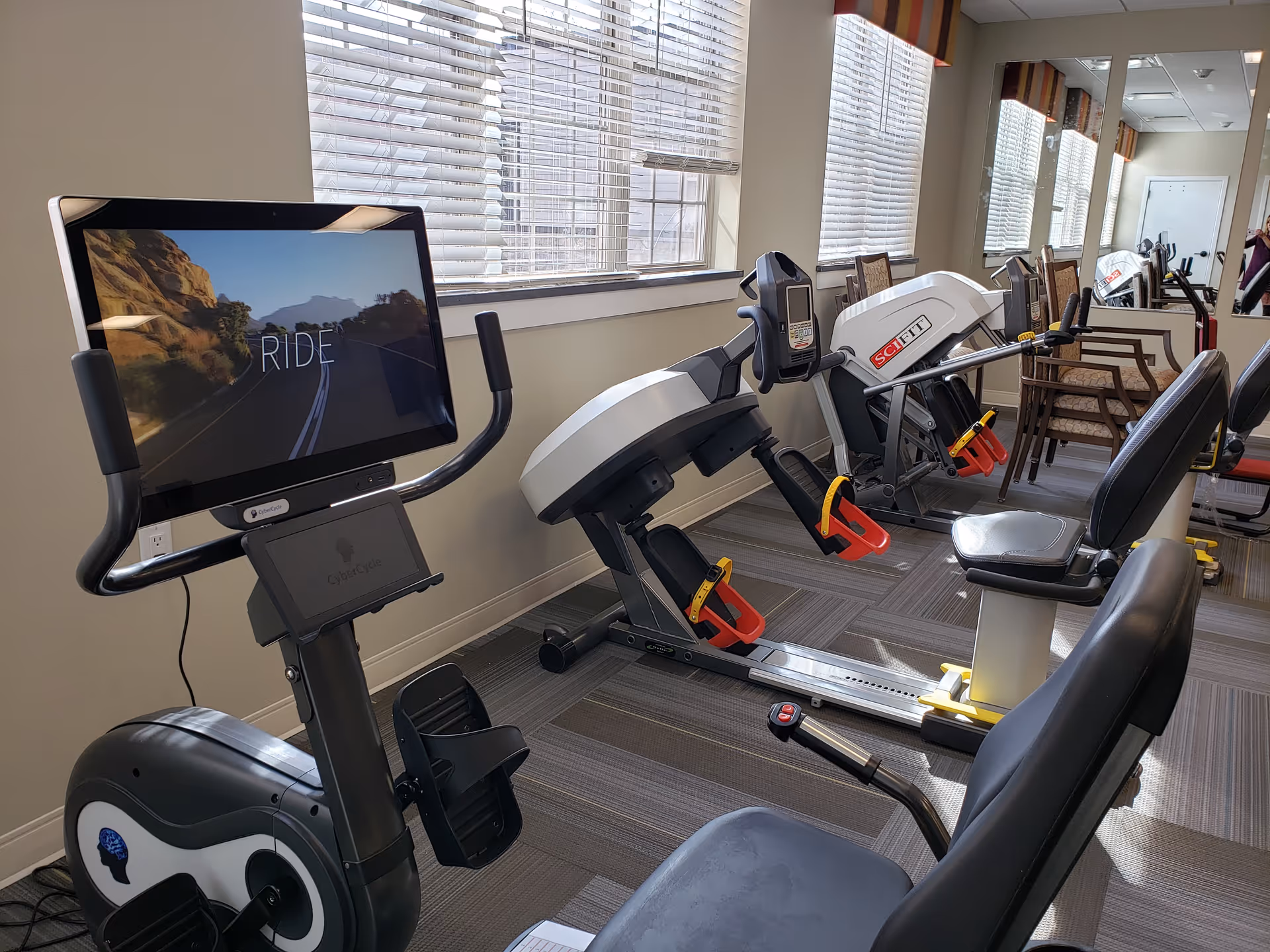Interior exercise room with stationary and recumbent bikes lined up by windows in a senior living facility.