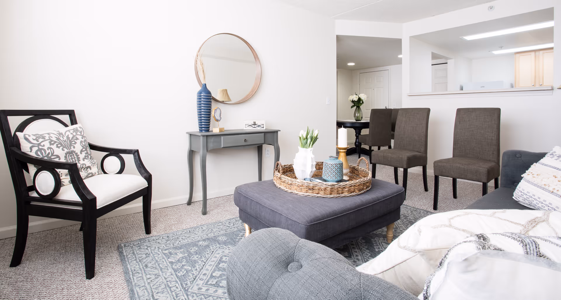 A bright and cozy living room with a gray upholstered ottoman topped with a wicker tray holding decorative items, a black wooden armchair with a patterned cushion, a small gray console table with a blue vase and a round mirror above it, and three brown dining chairs near a pass-through window to the kitchen.