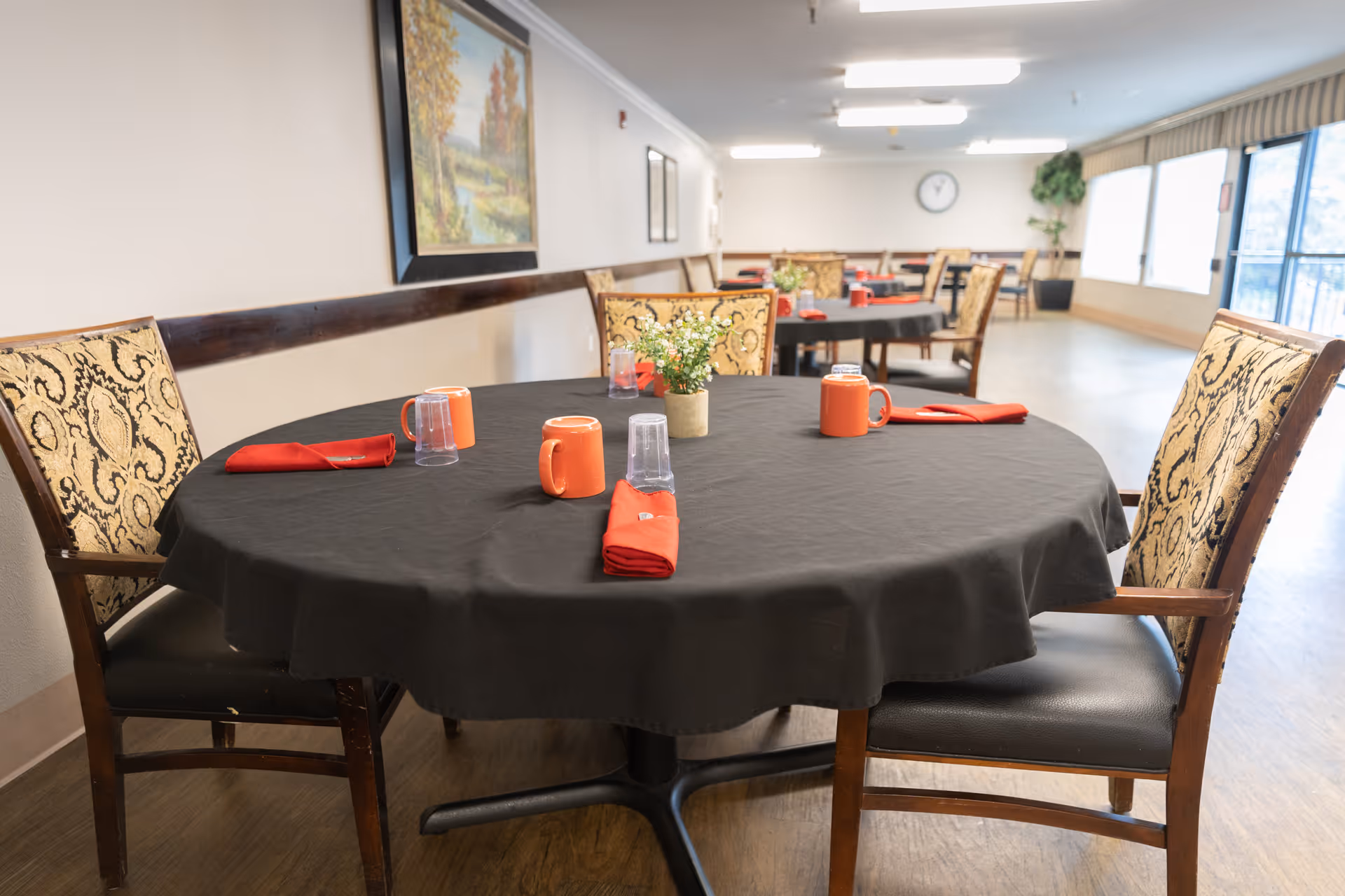 Round dining table with a black tablecloth set with orange mugs, red napkins and chairs in a bright memory-care dining room.