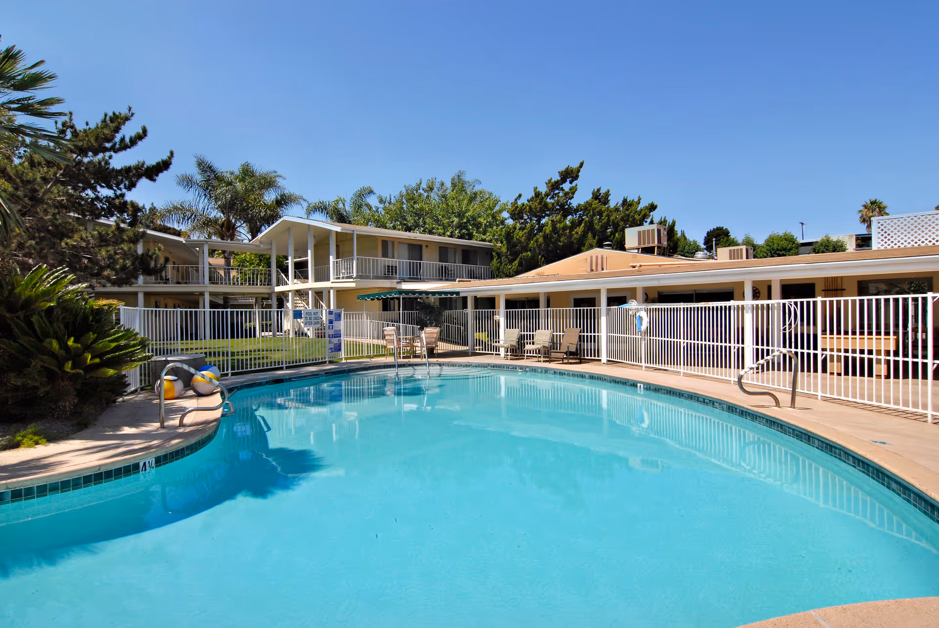 Outdoor swimming pool area at a senior living facility with clear blue water, surrounded by a white safety fence. There are lounge chairs and tables with umbrellas on the pool deck. In the background, there are two-story buildings with balconies and lush green trees under a clear blue sky.
