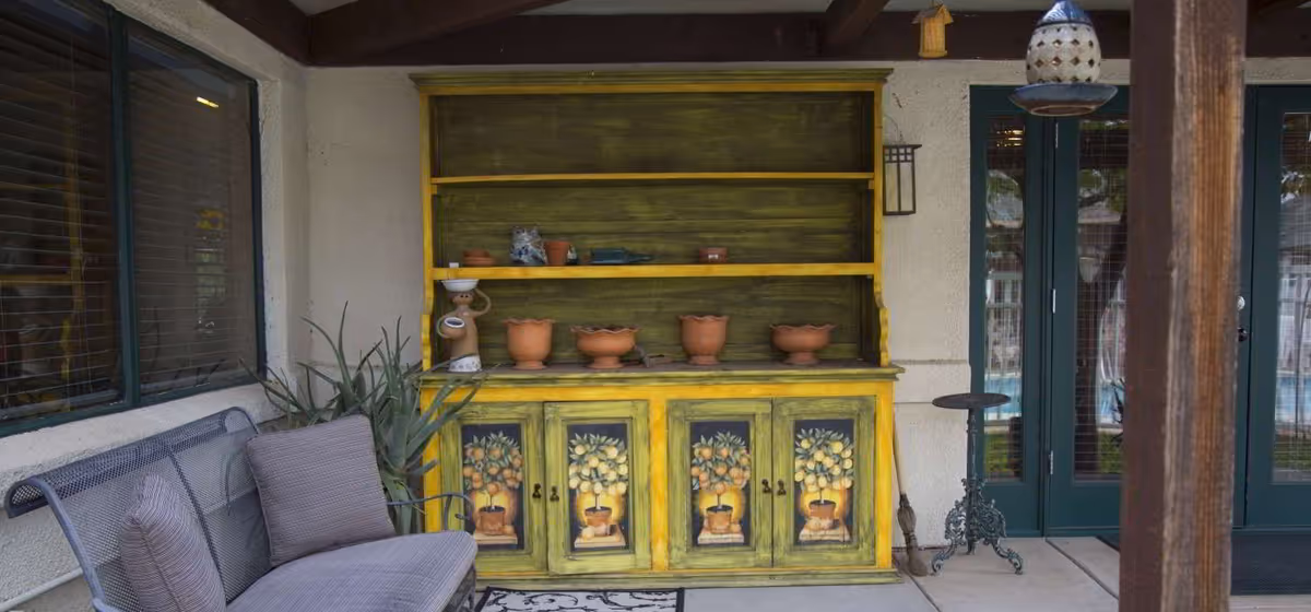Outdoor patio area with a yellow and green painted wooden cabinet featuring fruit designs, several terracotta pots on the shelves, a metal bench with gray cushions, a potted plant, a small round metal table, and green double doors with glass panes in the background.