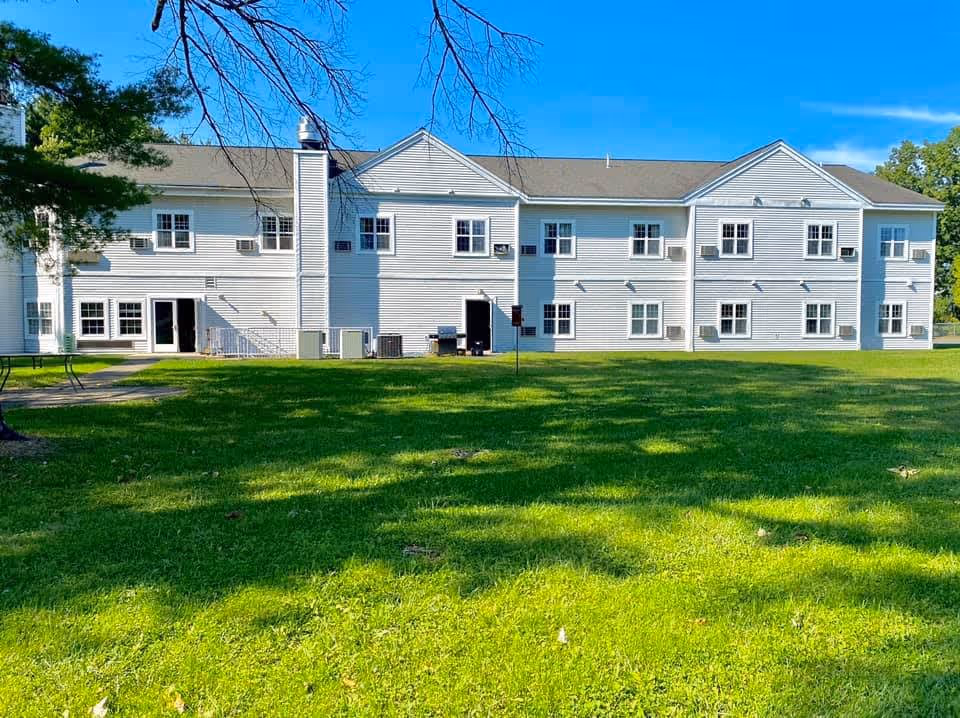 Exterior view of a two-story white building with multiple windows and a chimney, surrounded by a green lawn and trees under a clear blue sky.