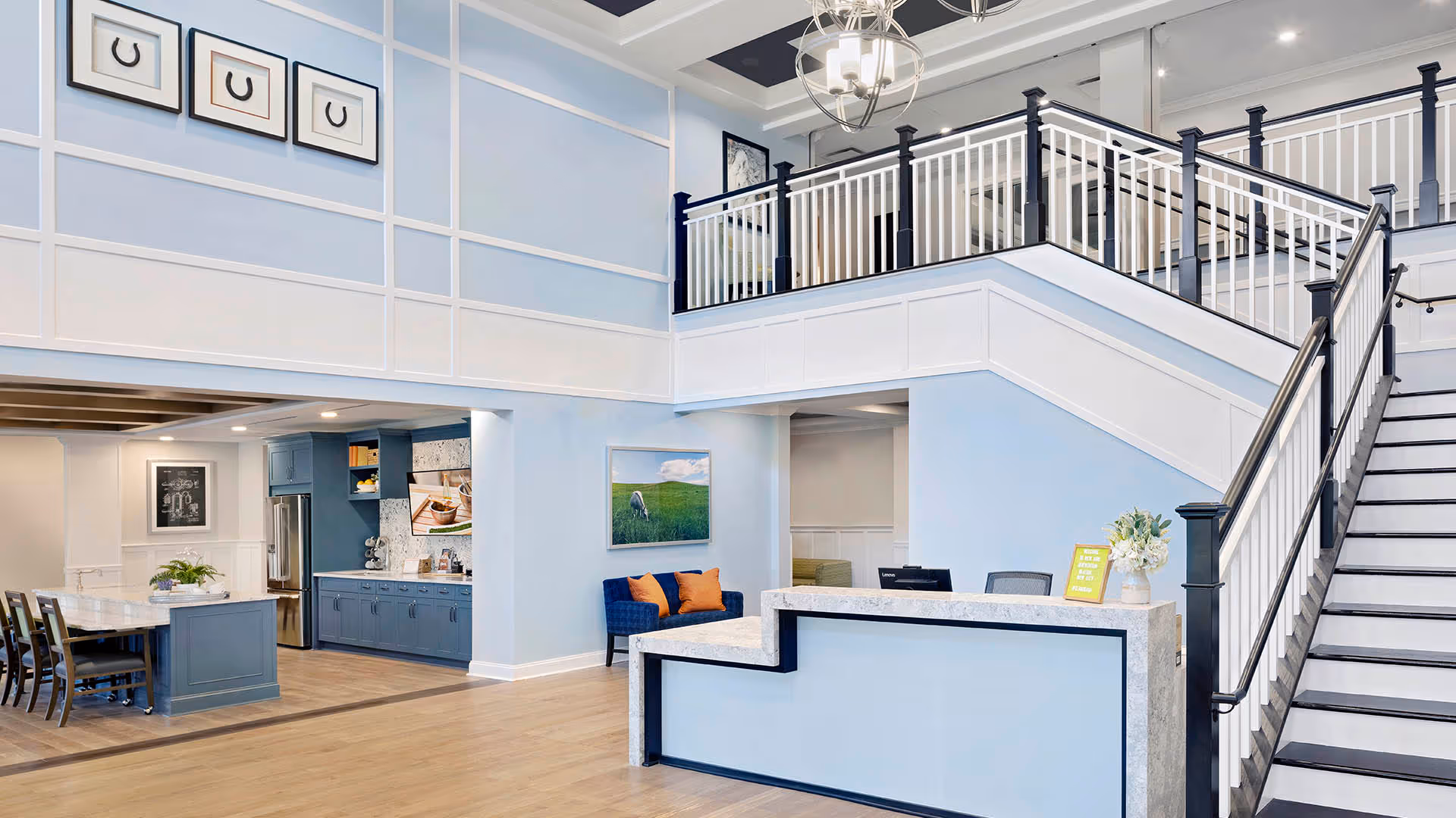Bright and spacious interior of a senior living facility with a modern reception desk in the foreground, a staircase with white railings leading to an upper level, and a kitchen area with blue cabinetry and a dining table with chairs in the background. The walls are painted light blue with white trim, and there are framed pictures and a chandelier hanging from the ceiling.