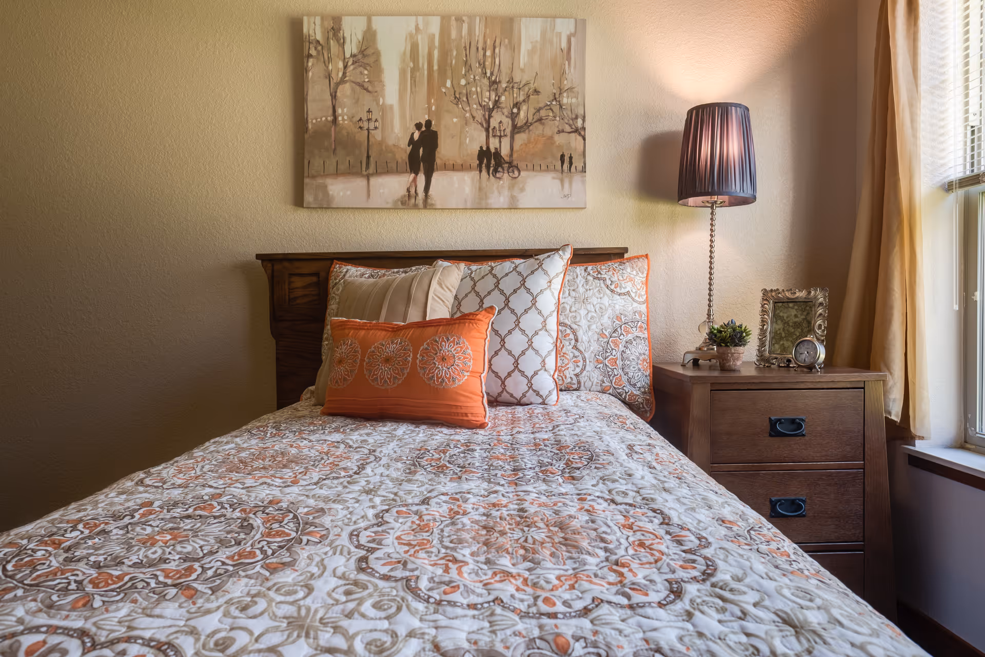 A decorated single bed with orange-and-white patterned bedding and pillows beside a wooden nightstand holding a lamp, small decor and a window.