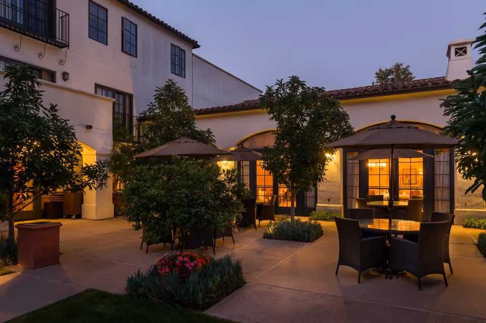 Outdoor courtyard area at dusk with several round tables and chairs under large umbrellas, surrounded by small trees and plants. The building in the background has large windows with warm interior lighting visible.
