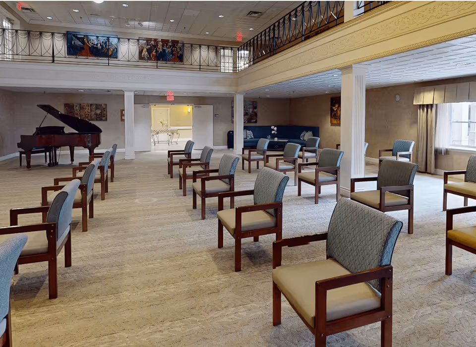 A spacious, well-lit common area with multiple wooden chairs arranged in rows facing a grand piano. The room has a high ceiling with a balcony railing above, decorative paintings on the walls, and large windows letting in natural light. There is also a seating area with a blue couch in the back corner.