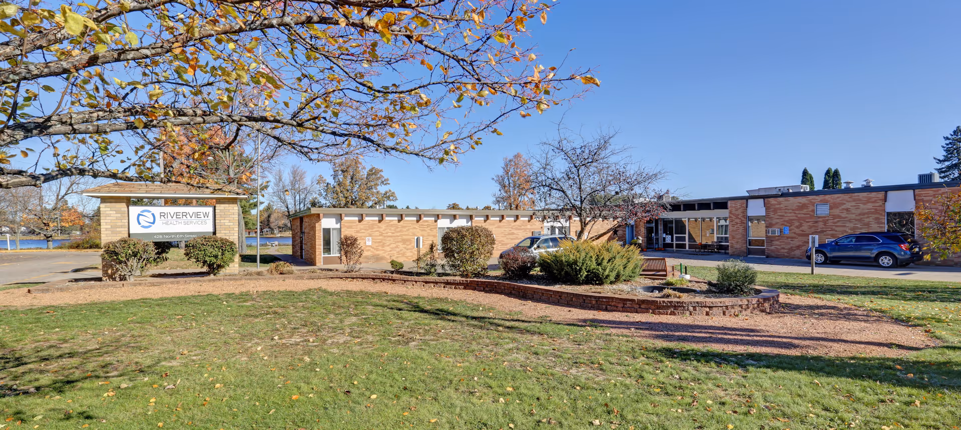 Exterior view of Riverview Health Services building with a lawn and landscaped area in the foreground, a sign with the facility name on the left, and a clear blue sky above.