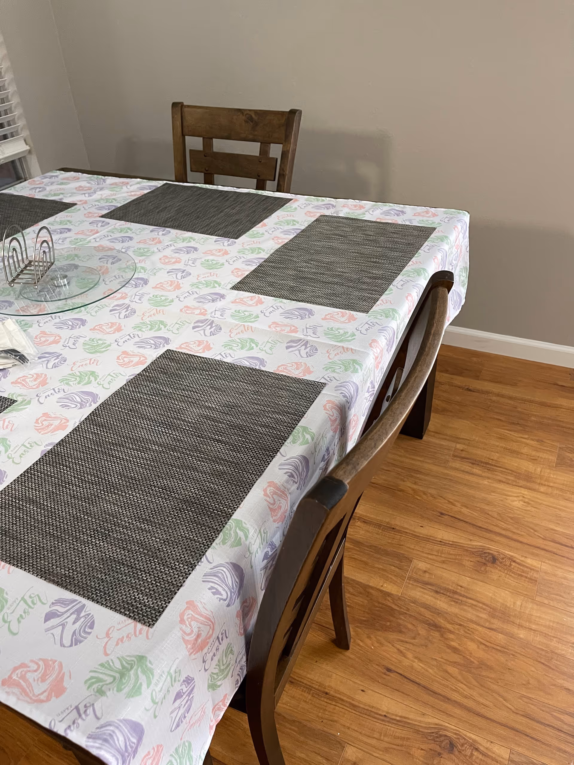 Dining table with a patterned tablecloth, dark placemats and wooden chairs on a hardwood floor in a dining area.