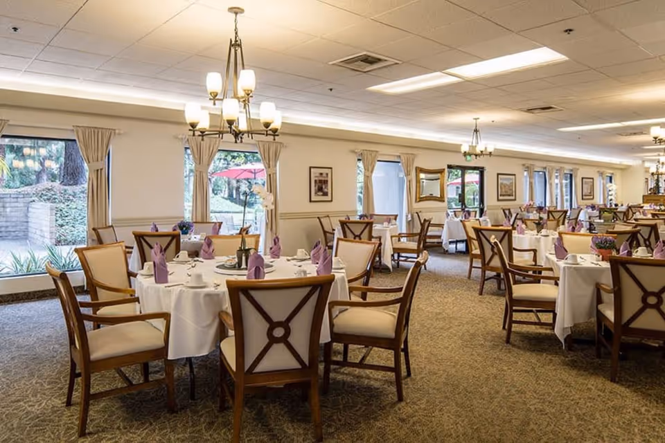 A spacious dining room with multiple round tables covered in white tablecloths, each set with purple folded napkins, cups, and plates. The room has large windows with beige curtains allowing natural light to enter, and elegant chandeliers hanging from the ceiling. Wooden chairs with cushioned seats surround the tables, and there are framed pictures and mirrors on the walls. Outside the windows, greenery and red umbrellas are visible.