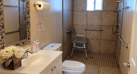 Accessible bathroom with sink, toilet, mirror, flowers, grab bars, and a shower chair.