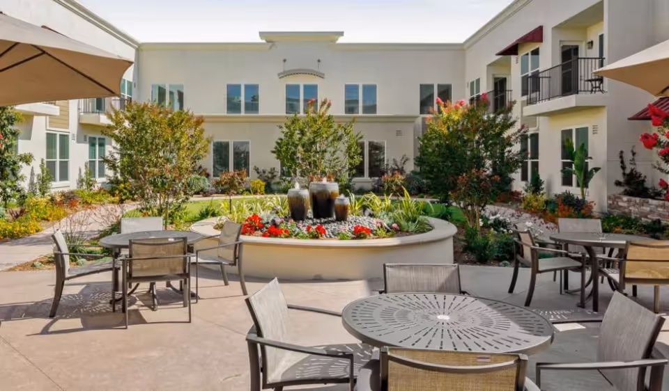 Outdoor courtyard area at Oakmont of Torrance featuring round metal tables with chairs, large umbrellas providing shade, a central circular planter with a water fountain, colorful flowers, and surrounding greenery. The courtyard is enclosed by a two-story building with multiple windows and balconies.