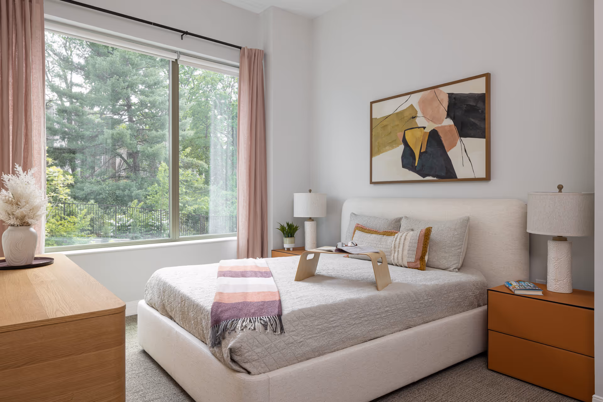 A bright bedroom with a large window showing green trees outside. The room features a neatly made bed with a light gray quilt, decorative pillows, and a striped throw blanket. There is a wooden tray on the bed, two bedside tables with matching lamps, a wooden dresser with a vase of dried flowers, and abstract artwork hanging above the bed.