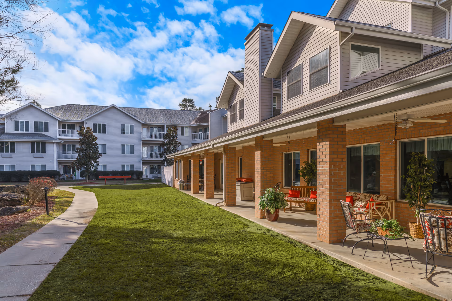 Outdoor courtyard and covered patio of a senior living facility with seating, lawn, and multi-story residences under a blue sky.