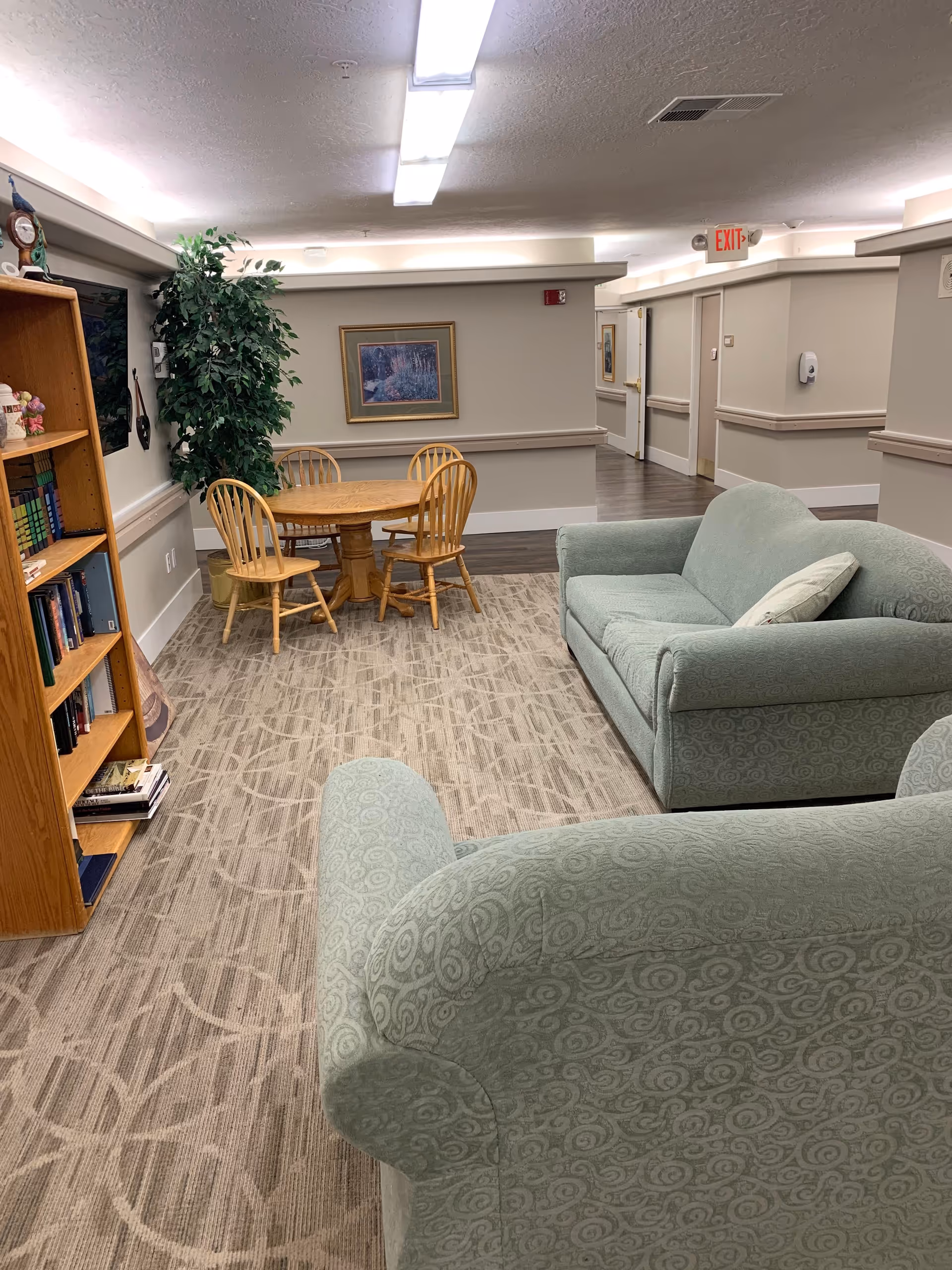 A cozy common area in an assisted living facility featuring two light green patterned sofas, a wooden round table with four matching chairs, a tall green plant in a pot, a wooden bookshelf filled with books, and framed artwork on the wall. The space has beige walls, patterned carpet, and overhead fluorescent lighting.