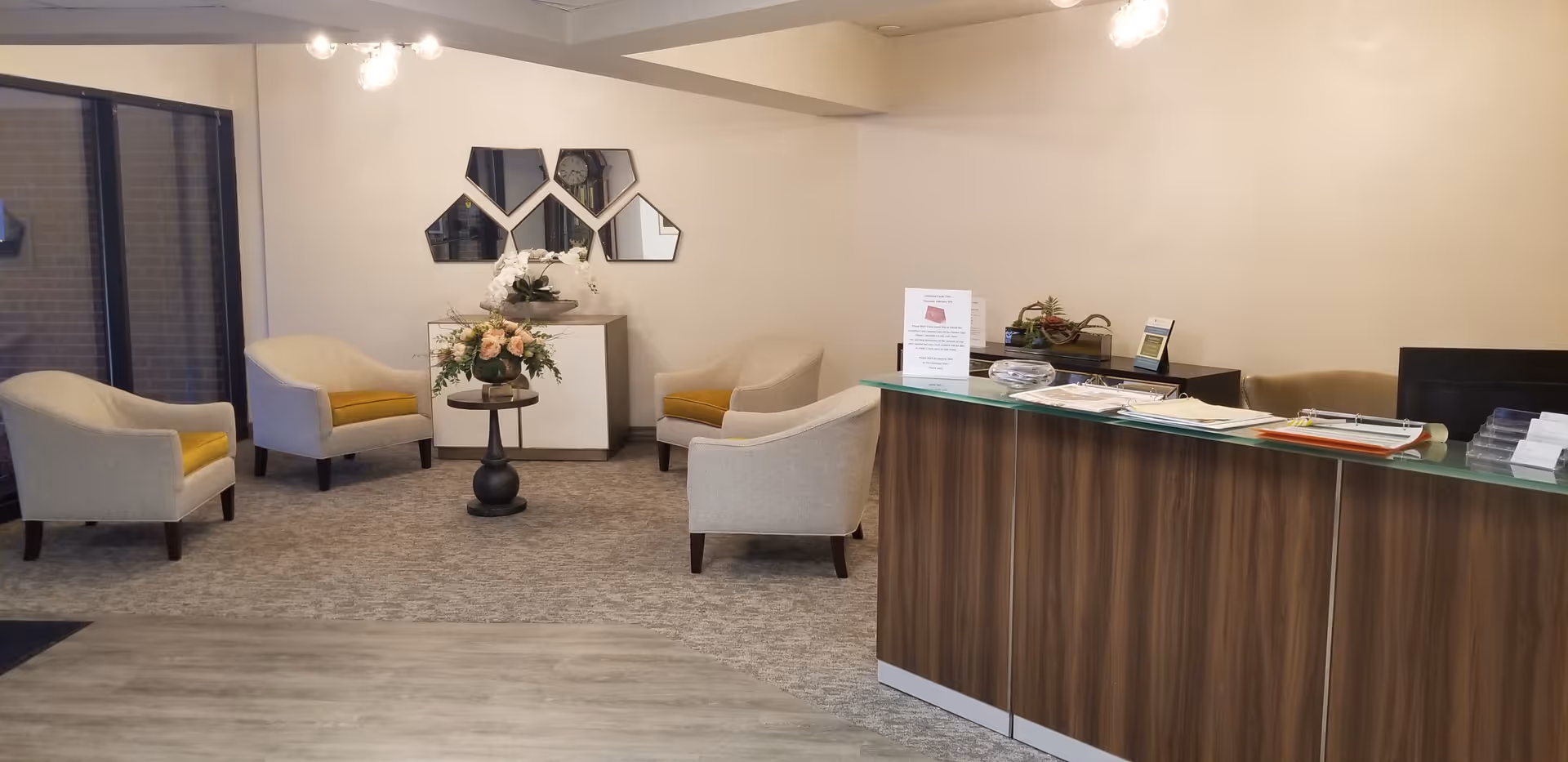 A senior living facility reception area with a wooden front desk on the right side, a small seating area with four beige armchairs with yellow cushions arranged around a small round table with a flower arrangement, and a cabinet with decorative items and geometric mirrors on the wall behind it.