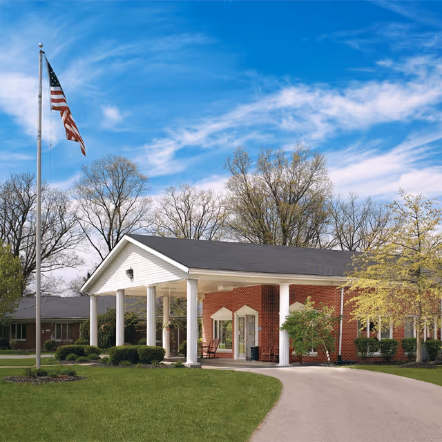 Brick senior living facility entrance with a white columned portico, driveway, American flag, and lawn under a blue sky.