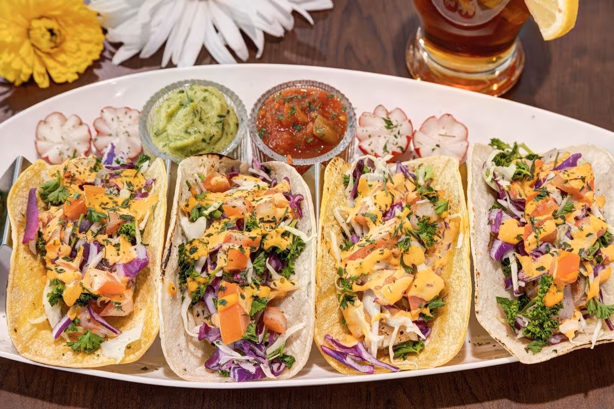 A white rectangular plate with four soft tacos filled with shredded cabbage, diced tomatoes, chopped parsley, and drizzled with a creamy orange sauce. The plate also has two small glass bowls, one with guacamole and the other with salsa, and garnished with thinly sliced radishes. A glass of iced tea with a lemon wedge is visible in the background on a wooden table.