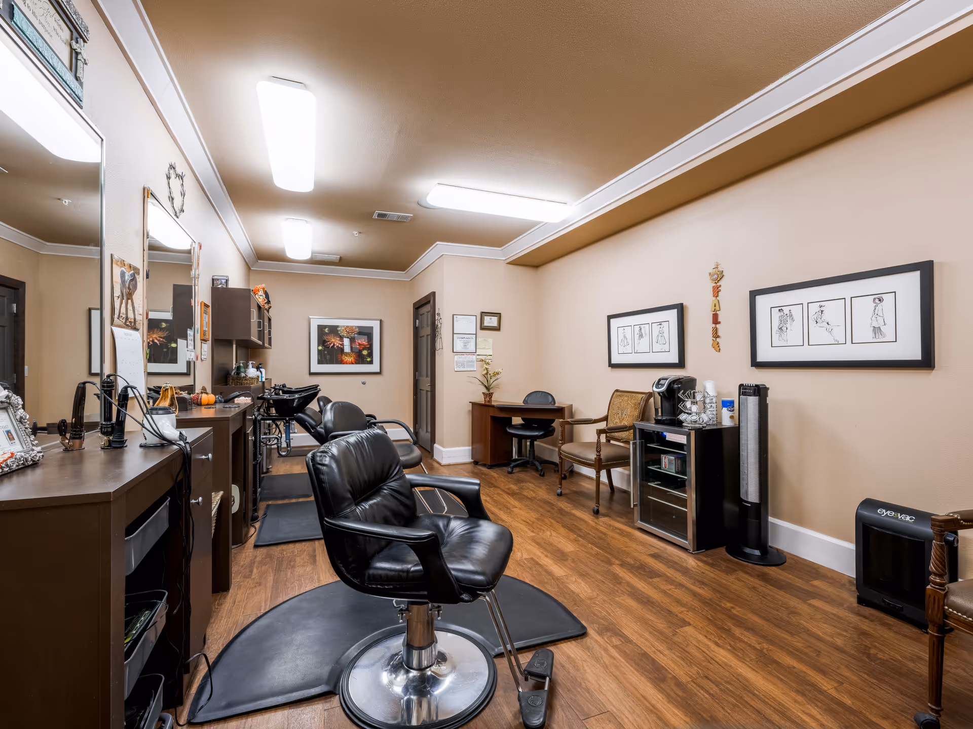 Interior view of a salon area in a senior living facility with black salon chairs, mirrors, wooden flooring, and beige walls decorated with framed artwork. There is a small desk with a chair, a coffee machine, and a fan on the right side of the room.