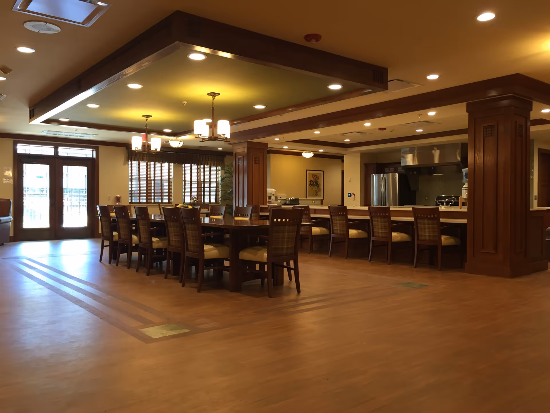 A spacious dining area with a long wooden table surrounded by chairs, a kitchen counter with stools, warm lighting fixtures on the ceiling, wooden flooring, and large windows with blinds letting in natural light.