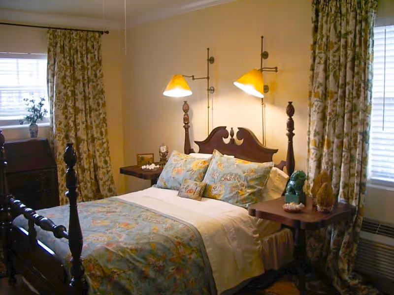 A cozy bedroom with a wooden bed frame featuring a floral patterned bedspread and matching pillows. The room has two wall-mounted lamps above the headboard, floral curtains on two windows, and small bedside tables with decorative items.