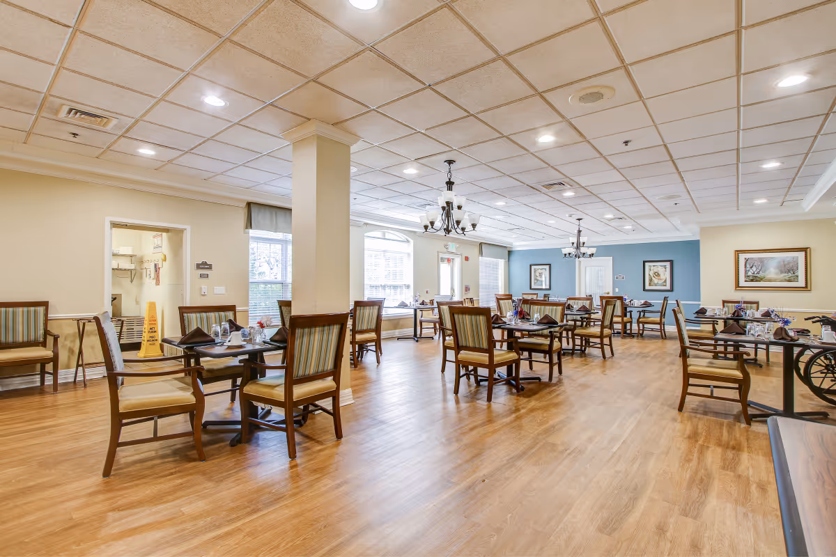 A spacious dining room in a senior living facility with multiple wooden tables and chairs arranged neatly. The room has large windows allowing natural light, light-colored walls with framed artwork, and chandeliers hanging from a tiled ceiling. The floor is wooden, and there is a wet floor caution sign near an open doorway.