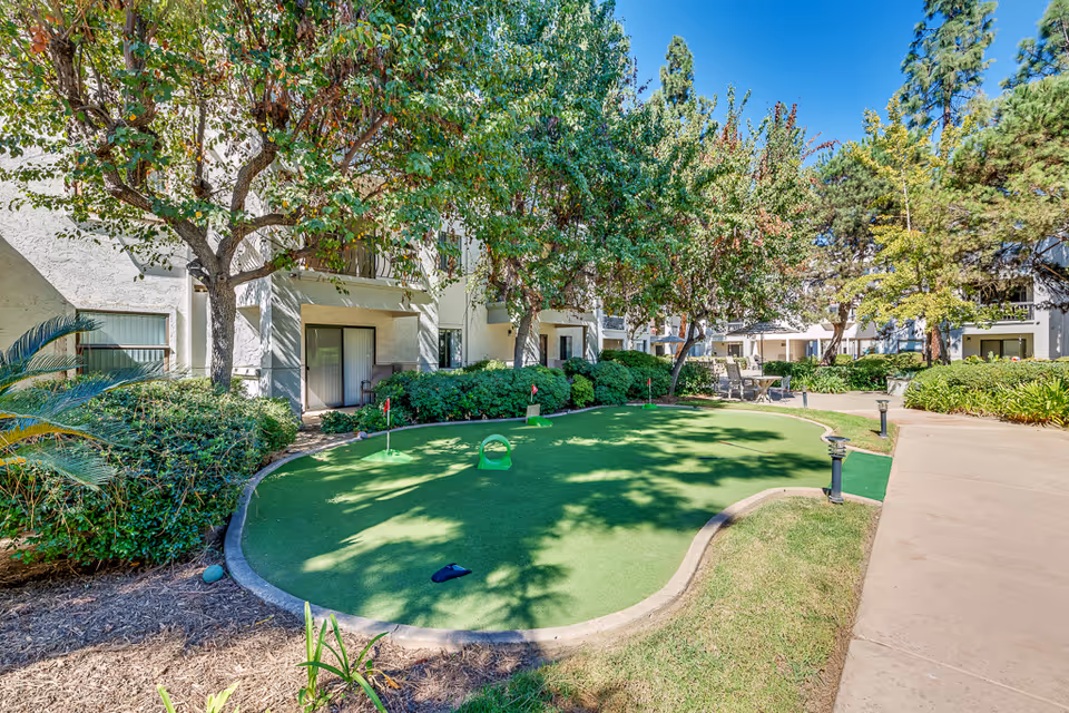 Outdoor putting green surrounded by trees and bushes at a senior living facility, with a paved walkway and residential building in the background under a clear blue sky.