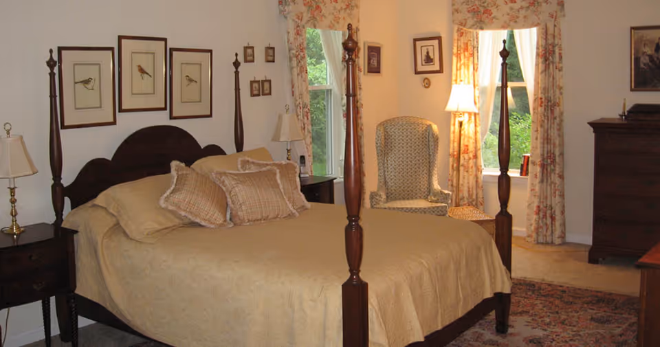 A cozy bedroom with a large wooden four-poster bed covered in beige bedding and multiple pillows. The room features two bedside tables with lamps, framed bird artwork on the wall above the bed, floral curtains on the windows, a patterned armchair near the window, a tall wooden dresser, and a floor lamp providing warm lighting.