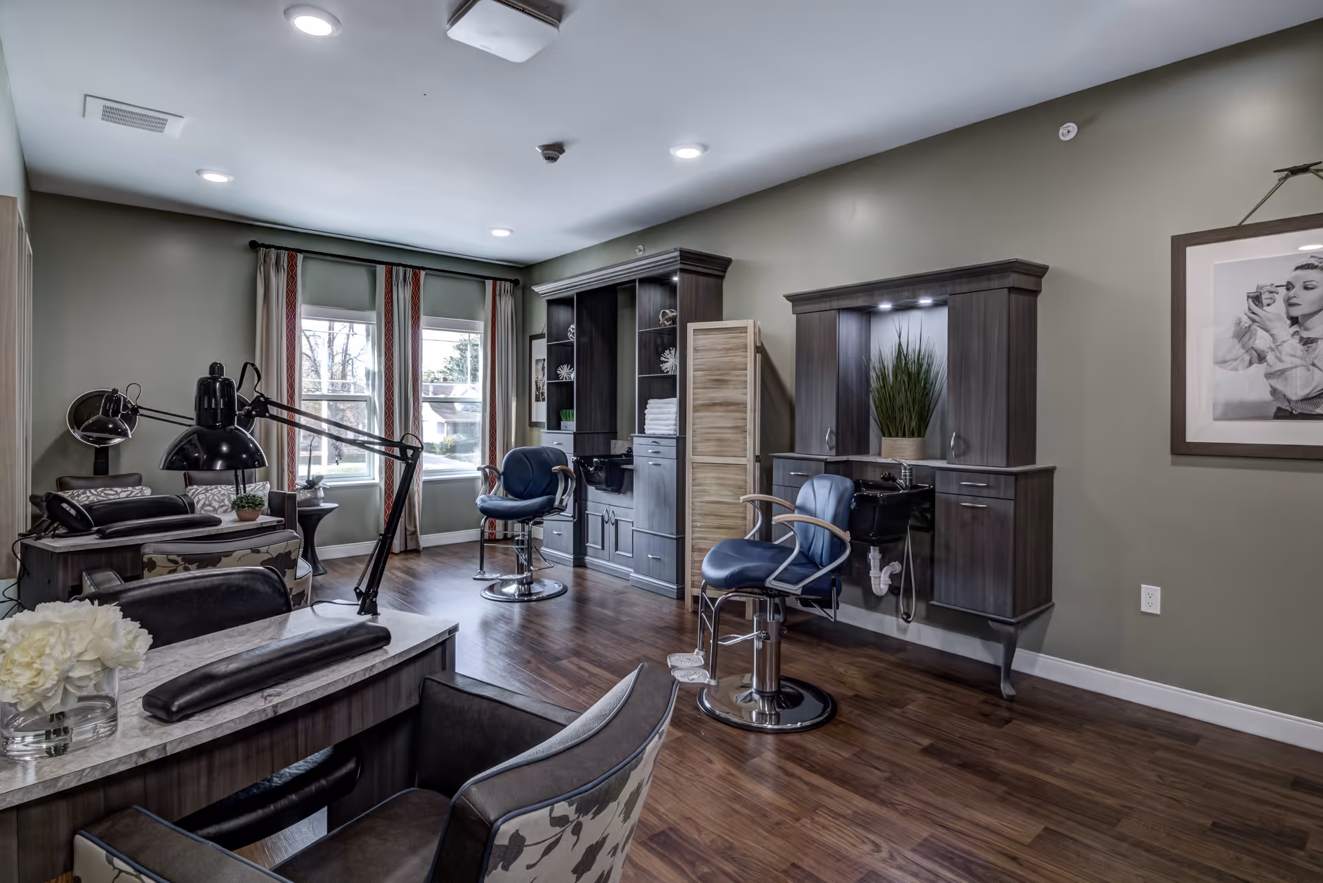 A well-lit salon area in an assisted living facility featuring two blue salon chairs in front of dark wood cabinetry with sinks, a manicure table with a black lamp and cushioned chairs, and a window with curtains letting in natural light. The room has wooden flooring and neutral-colored walls with a framed black and white photo on one wall.