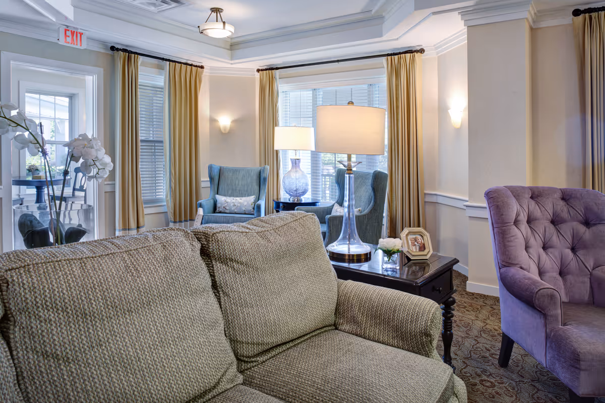 A cozy living room area with a green textured sofa in the foreground, a purple tufted armchair to the right, and two blue armchairs near the windows with yellow curtains. A side table with a clear glass lamp, a small flower arrangement, and a framed photo is placed between the sofa and armchairs. The room has soft lighting and a patterned carpet.