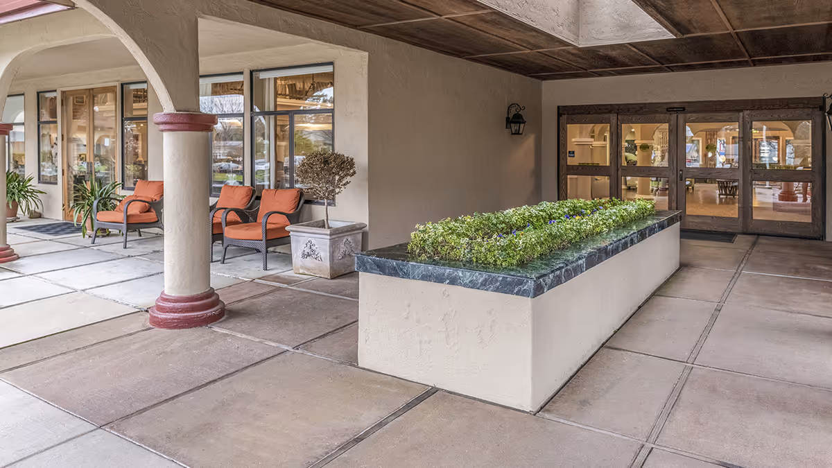 Covered outdoor seating area with two orange cushioned chairs, potted plants, a rectangular planter with green foliage, and glass doors leading inside a building.