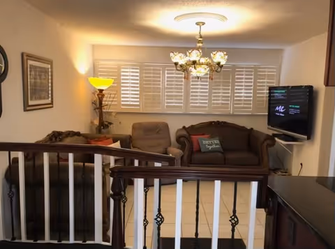 Cozy living room with two sofas, a recliner, a chandelier, wall-mounted TV, and a railing in the foreground.